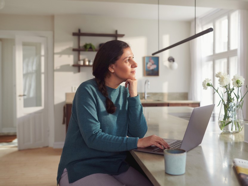 Kvinne sitter med en tekopp og en laptop på kjøkkenet og ser ut i luften og tenker. Foto.