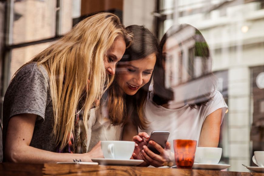 Tre damer ser på mobilen på en kafe.